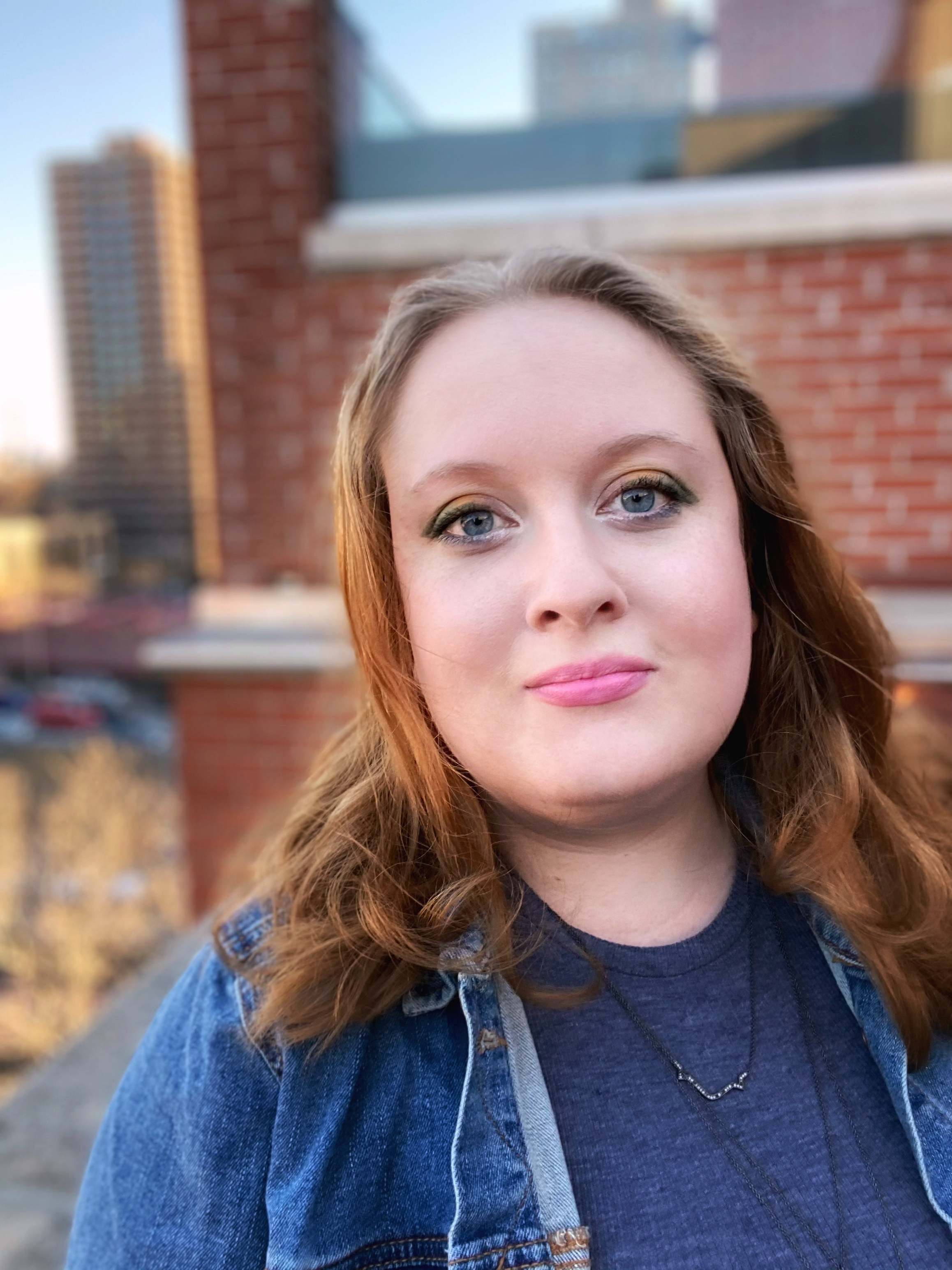 Image of founder Savvy Gatton. White woman with mid-length red wavy hair, blue eyes, and a round face. She is wearing a blue shirt, denim jacket, and black layered necklace. 